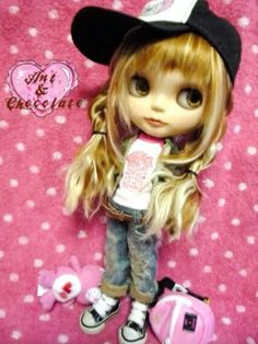 a doll with blonde hair wearing a hat and holding a pink bag on a bed