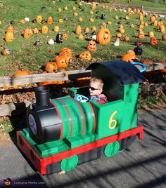 a small child in a green and red train with pumpkins on the field behind him