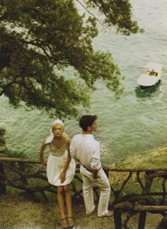 a man and woman standing next to each other on a hillside near water with boats in the background