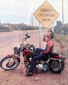 a man sitting on a motorcycle drinking from a water bottle next to a road sign