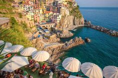 people are sitting at tables near the ocean with umbrellas and buildings in the background