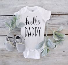 a baby bodysuit with the words hello daddy next to shoes and eucalyptus leaves on a wooden background