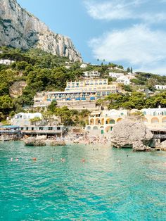 people are swimming in the blue water next to houses and cliffs on a hill side