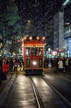 a red trolley car traveling down a street next to tall buildings with christmas lights on them