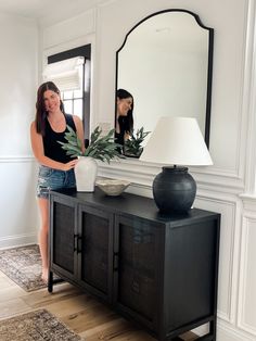 a woman standing in front of a mirror next to a table with a vase on it