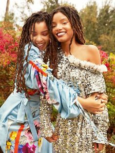 two women in sequins hugging each other and smiling at the camera with flowers behind them