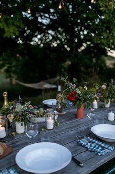 a wooden table topped with white plates and vases filled with flowers next to candles