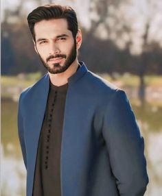 a man with a beard wearing a blue suit and black shirt standing in front of a body of water