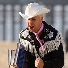 a man wearing a white cowboy hat and holding a book in his right hand while walking on the beach