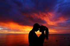 a man and woman are kissing in front of the ocean as the sun goes down