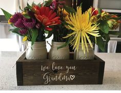 three mason jars with flowers in them sitting on a kitchen counter next to an i love you mom sign