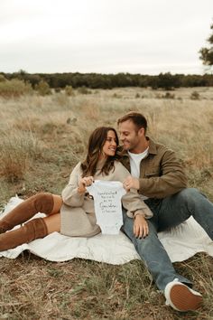 a man and woman sitting on top of a blanket in the grass holding a sign