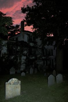 an old house with tombstones in the grass at night under a pink sky filled with clouds