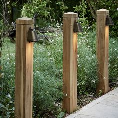 three wooden posts with lights on them in front of some bushes and trees, along side a sidewalk