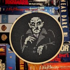 a close up of a embroidery on a wall with many books behind it and a cross - stitch picture of a man