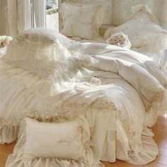 a white bed with ruffles and pillows on it in a room next to a window