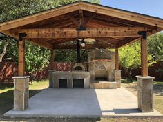 an outdoor grill area with stone pillars and a wooden roof, surrounded by grass and trees