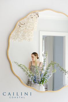 a woman standing in front of a mirror holding a vase with flowers on the table