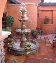 a fountain in the middle of a courtyard with potted plants on either side and an orange wall behind it