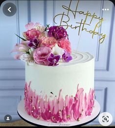 a white cake with pink and purple flowers on top that says happy birthday in gold lettering