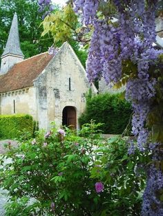 an old church surrounded by trees and bushes with purple flowers in the foreground on a sunny day