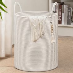 a large white basket with tassels on the bottom and handles is shown in front of a bookshelf