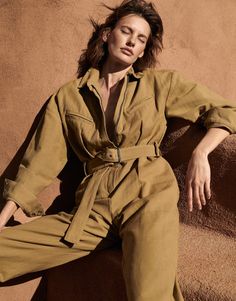 a woman sitting on top of a rock next to a brown wall with her hair blowing in the wind