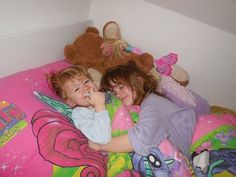 two young children laying in bed with stuffed animals on the wall behind them and smiling at the camera