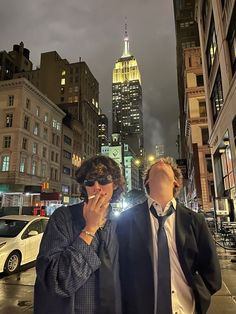 two men standing next to each other in front of tall buildings with lights on them