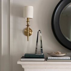 a white mantle with books and a lamp on it in front of a round mirror