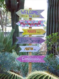 a wooden sign with many signs on it in front of cactus plants and trees,