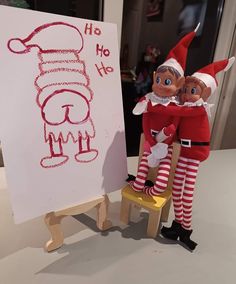 two elfs are standing next to a drawing on a easel and one is sitting on a stool