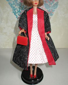 a doll is wearing a red and black coat with polka dots on the collar, white dress, and red shoes