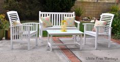 three white chairs and a table with pillows on it in a backyard area next to some plants