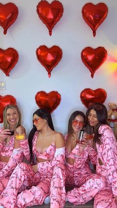 three women in pink pajamas sitting on a bed with red heart balloons hanging above them