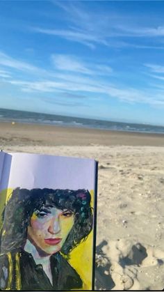 an open book sitting on top of a sandy beach next to the ocean with a painting of a woman's face