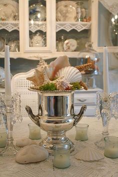 a silver vase filled with seashells on top of a white table cloth next to candles