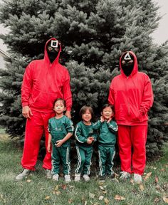 three children wearing red and green outfits standing in front of a christmas tree with two ninjas on their heads