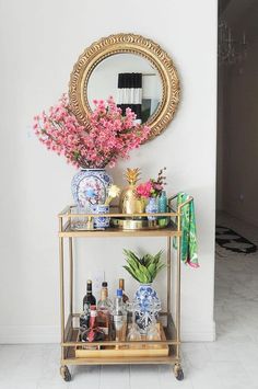 a gold bar cart filled with bottles and vases next to a mirror on the wall