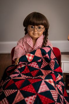 a doll is sitting in a chair with a quilt on it's lap and wearing glasses