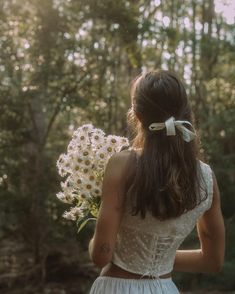 a woman in a white dress holding a bouquet of flowers and looking at the woods