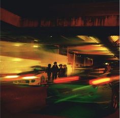 blurry photograph of people standing in front of a building at night with cars passing by