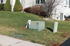 an electrical box sitting in the grass next to a trash can and garbage can on a residential street