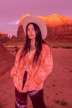 a woman standing in the desert wearing a cowboy hat and an orange shirt with purple tie dye