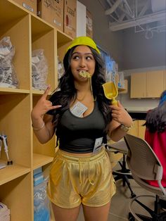 a woman in yellow shorts and a black top is eating something with her hands while standing next to shelves