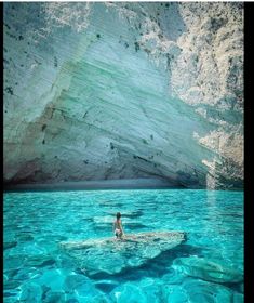 a person is swimming in the blue water near a large rock formation with a mountain behind them