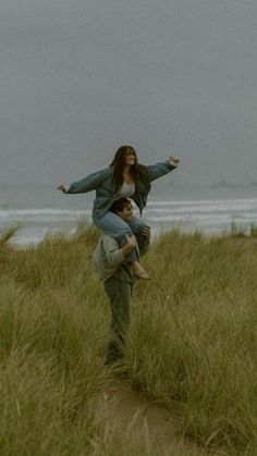 two people standing in tall grass near the ocean with one person on his back and another holding an umbrella