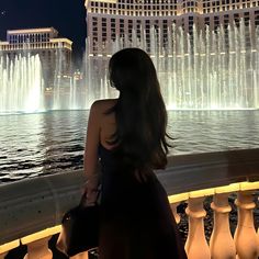 a woman standing in front of a fountain at night with the city lights behind her