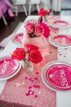 the table is set with pink and silver plates, napkins, and red roses