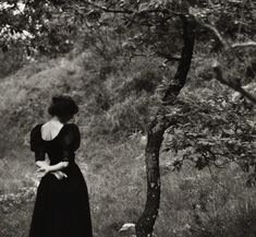 a woman in a black dress standing next to a tree with her hands on her hips